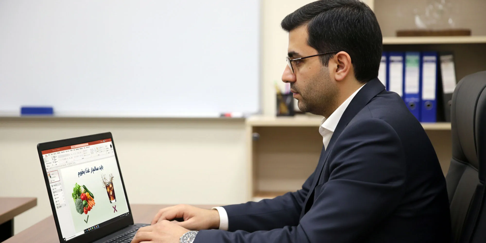 مردی کت و شلوارپوش با عینک، در یک دفتر کار، در حال کار با لپ‌تاپ دیده می‌شود. صفحه لپ‌تاپ او یک اسلاید پاورپوینت را نمایش می‌دهد که به فارسی می‌گوید "باید سالم‌تر غذا بخوریم" و یک مقایسه تصویری بین سبزیجات و نوشابه را نشان می‌دهد. پشت سر او یک تخته سفید و قفسه‌ای حاوی پرونده‌ها قرار دارد.