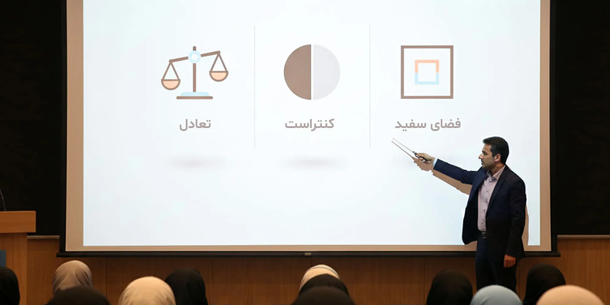 یک مرد در حال سخنرانی در مقابل یک صفحه نمایش است که سه اصل طراحی را به فارسی نشان می‌دهد: تعادل، کنتراست و فضای سفید.