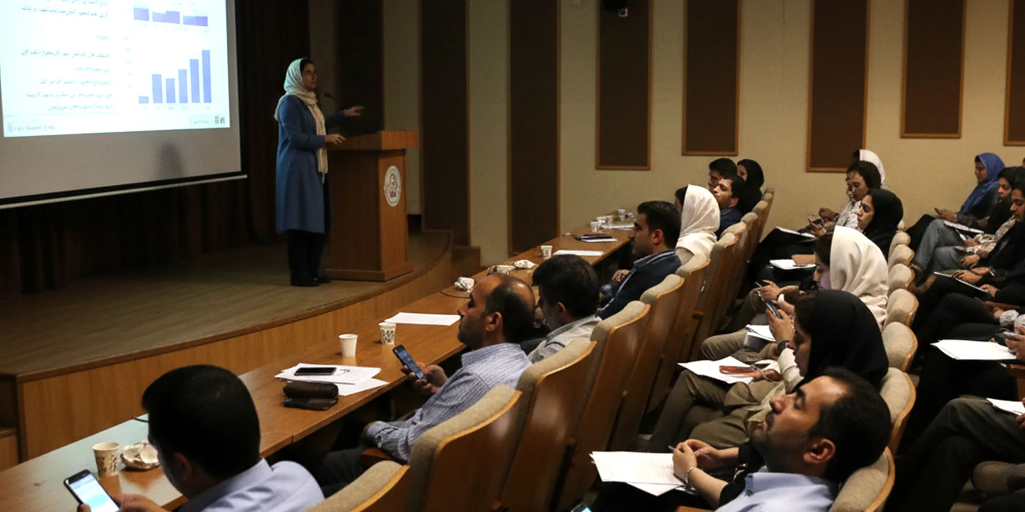 تصویر یک سالن سخنرانی را نشان می‌دهد. سخنران در حال ارائه مطالبی است که بر روی پرده نمایش است. به علت اینکه ارائه وی ریتم و کشش ندارد. مخاطبان یا به گوشی موبایل خود نگاه می کنند یا اینکه خوابشان برده است. 