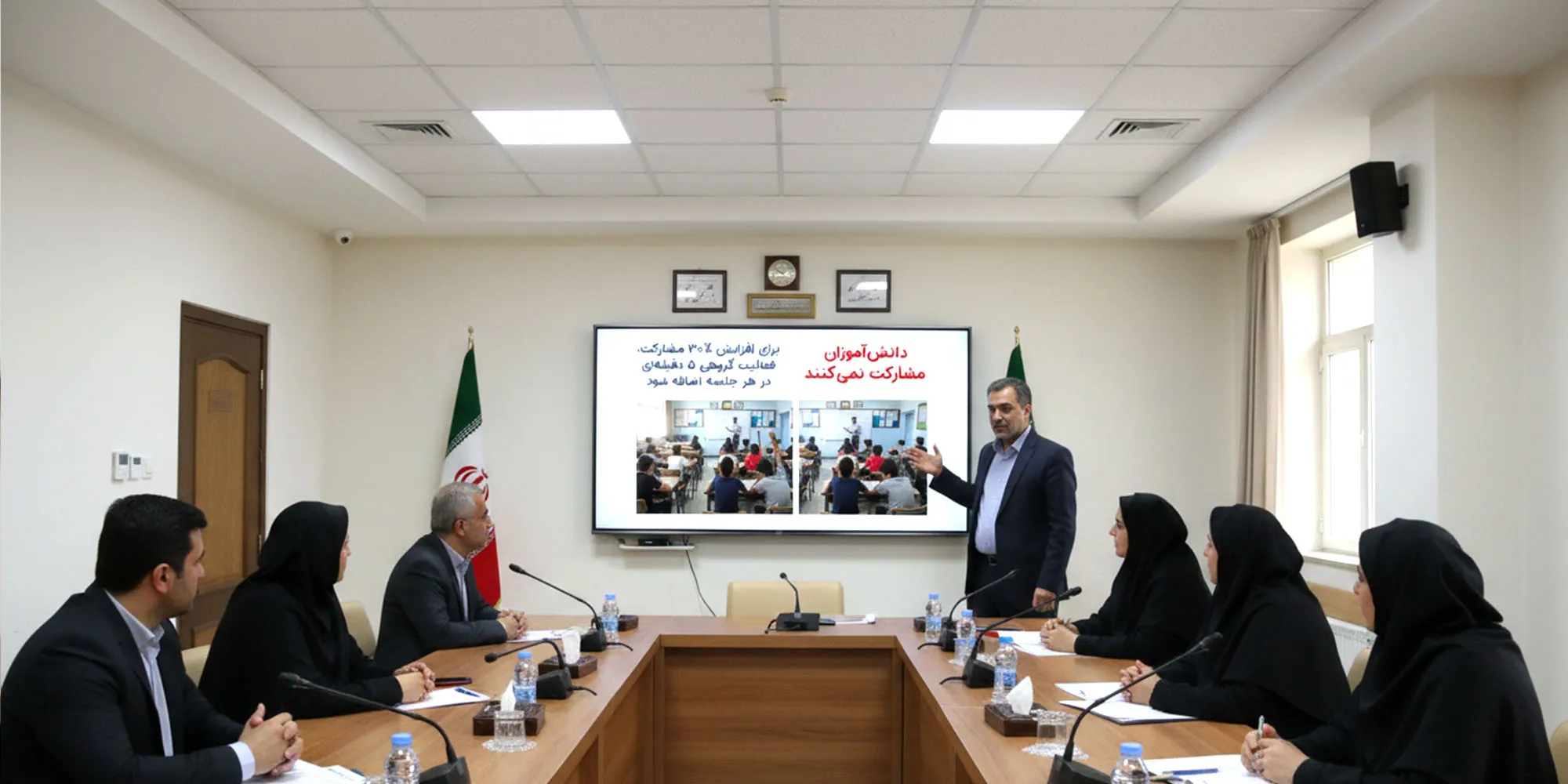 مردی در حال ارائه در یک جلسه رسمی با میز کنفرانس U-شکل است. هفت نفر دیگر، شامل پنج زن با حجاب، به او گوش می‌دهند. اسلاید روی صفحه نمایش پشت سر او حاوی متنی فارسی در مورد **"دانش‌آموزان مشارکت نمی‌کنند"** و دو تصویر مرتبط با فعالیت‌های کلاسی است. پرچم ایران نیز در کنار صفحه دیده می‌شود.
