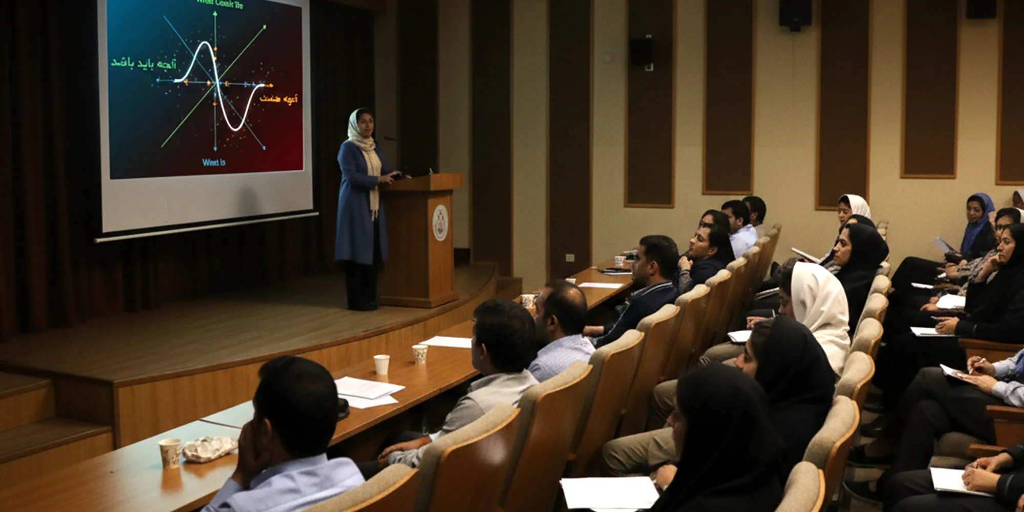  تصویر نمایی از یک سالن سخنرانی را نشان می‌دهد . سخنران در حال ارائه است و مخاطبان در حال گوش دادن به سخنان او هستند. 